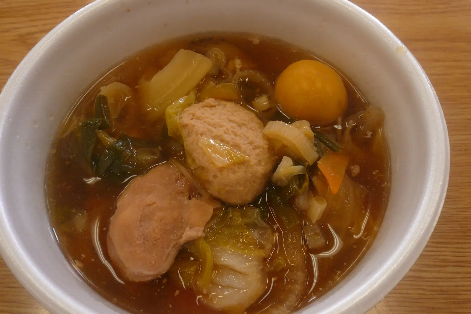 A white bowl of chankonabe stew holding a fish ball, an egg yolk, cabbage and carrot in a brown broth, on a wooden table
