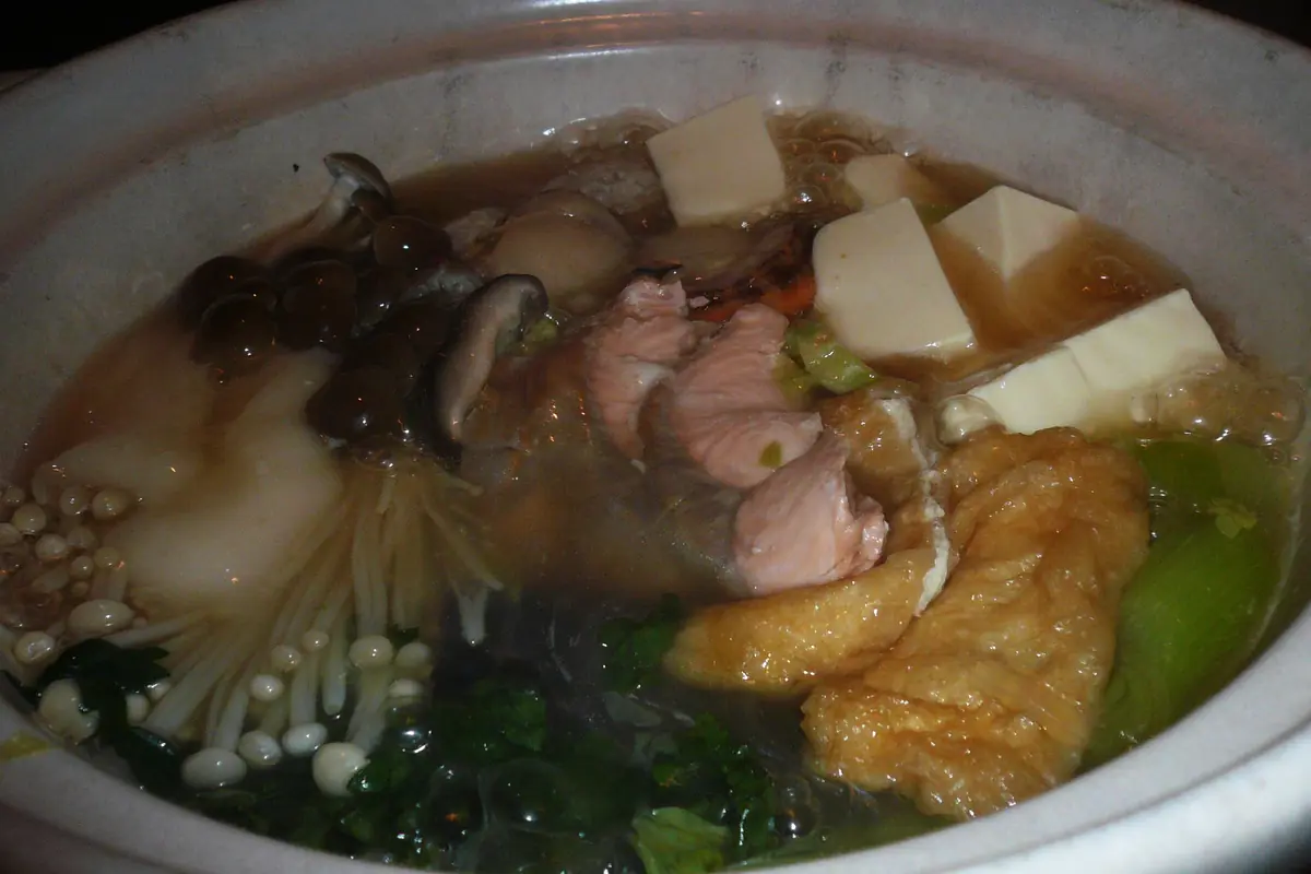 A clay pot of chankonabe stew simmering on a table, with sliced chicken, tofu, mushrooms, leafy greens and noodles in the broth