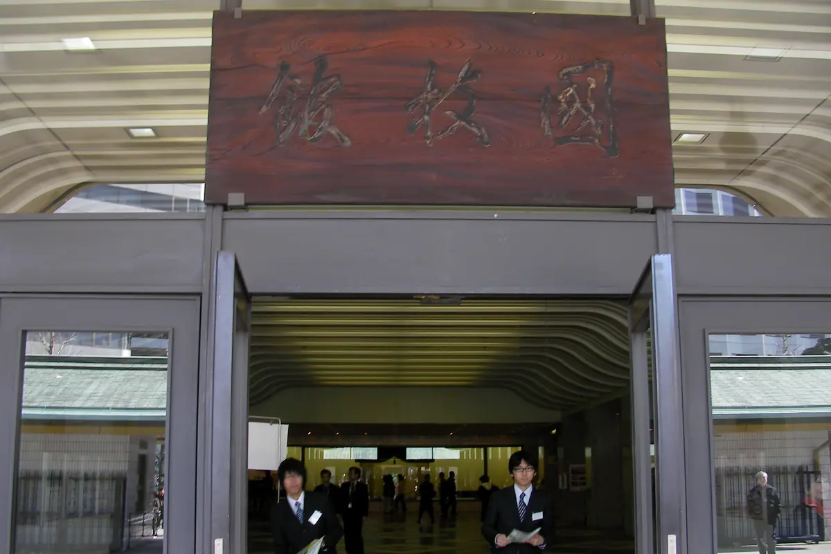 The wooden entrance sign and glass doors of the Ryogoku Kokugikan sumo arena, with two staff members standing in the doorway