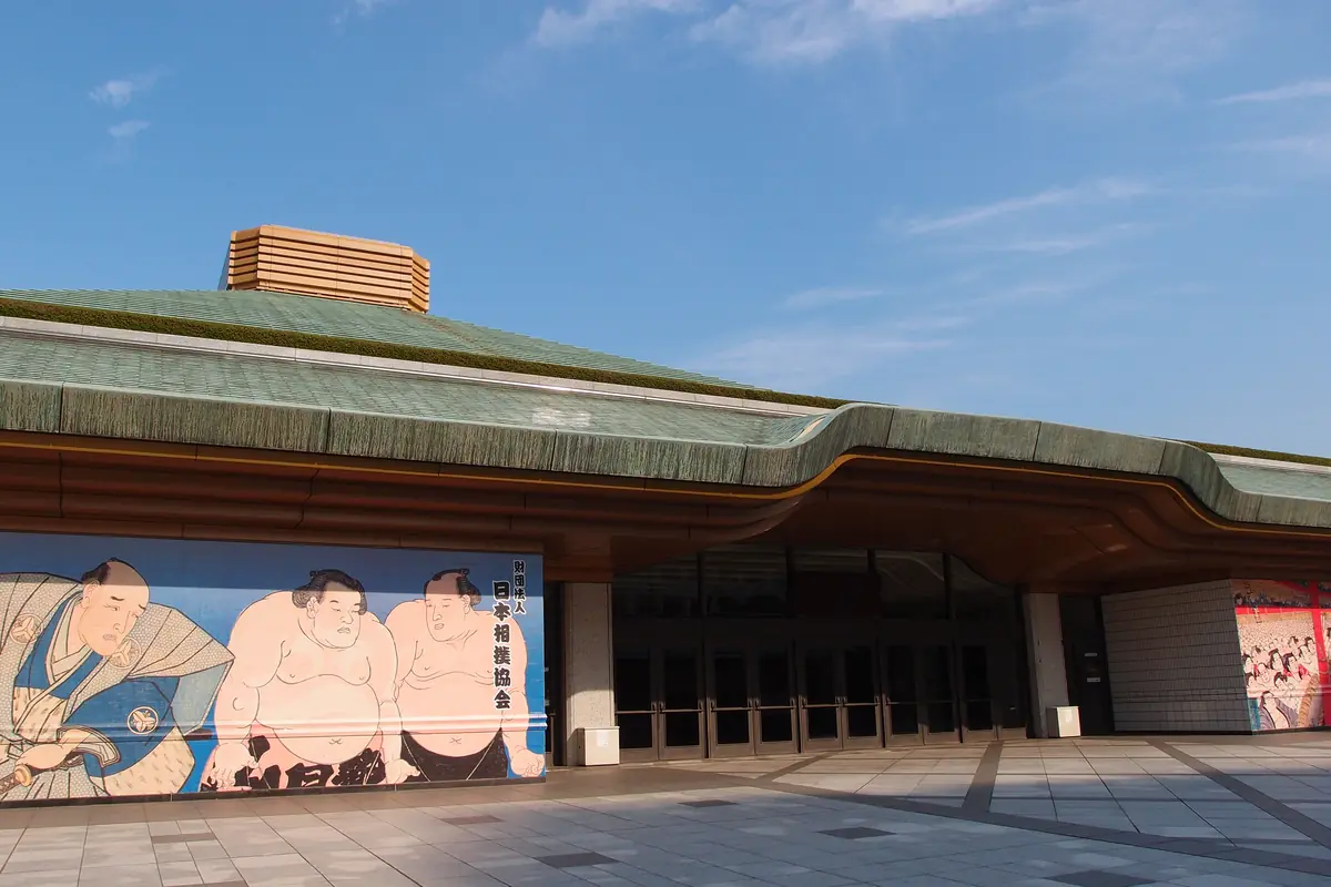 The long green roof and entrance hall of the Ryogoku Kokugikan sumo arena in Tokyo, with a painted mural of wrestlers on the wall beside the doors