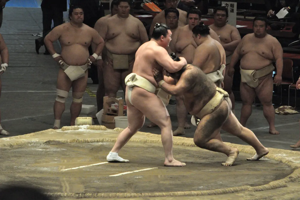 Two sumo wrestlers locked in a bout on a clay ring at a practice session, with other wrestlers watching from behind the ring and spectators in the stands