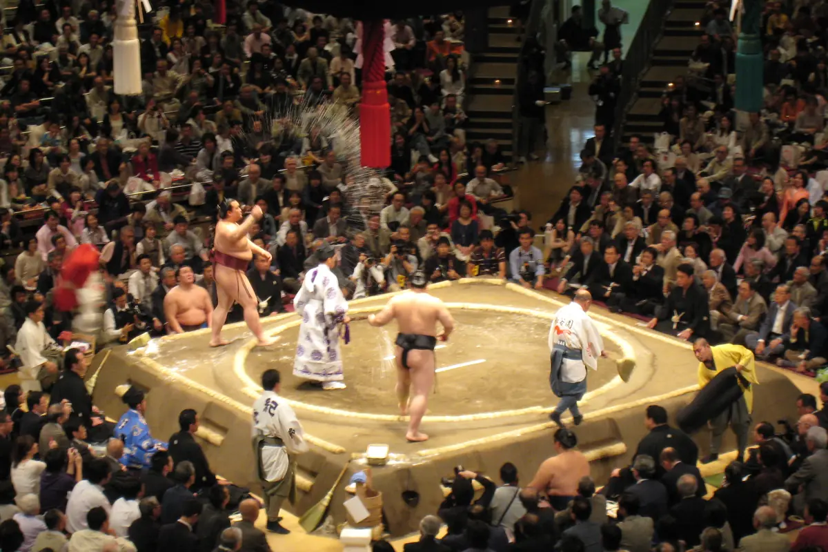 A sumo wrestler throwing salt into a raised clay ring in a packed arena, with a referee in white robes and other wrestlers beside the ring