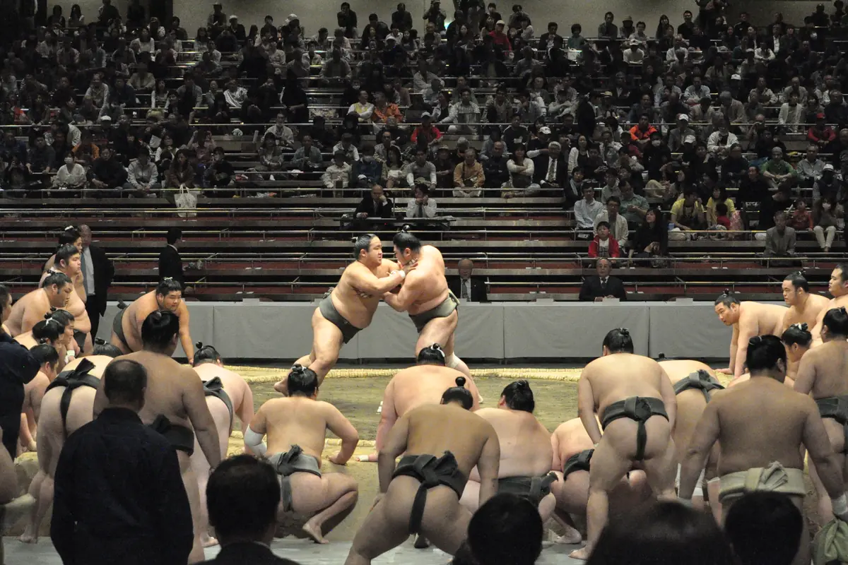 A crowd of sumo wrestlers crouching around a clay ring at a practice session, with two wrestlers in a bout at the centre and spectators in the stands behind