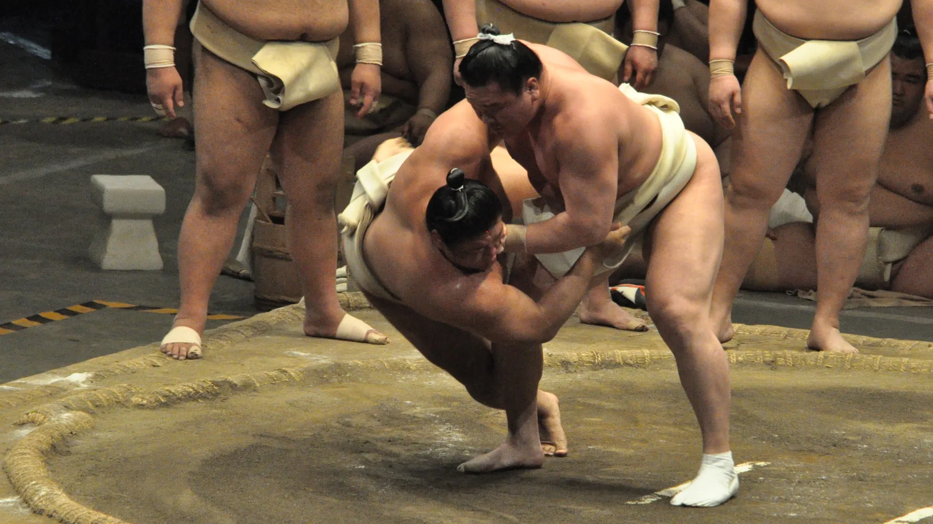 Two sumo wrestlers locked in a bout on a clay ring edged with rice-straw bales, with other wrestlers standing and kneeling behind them