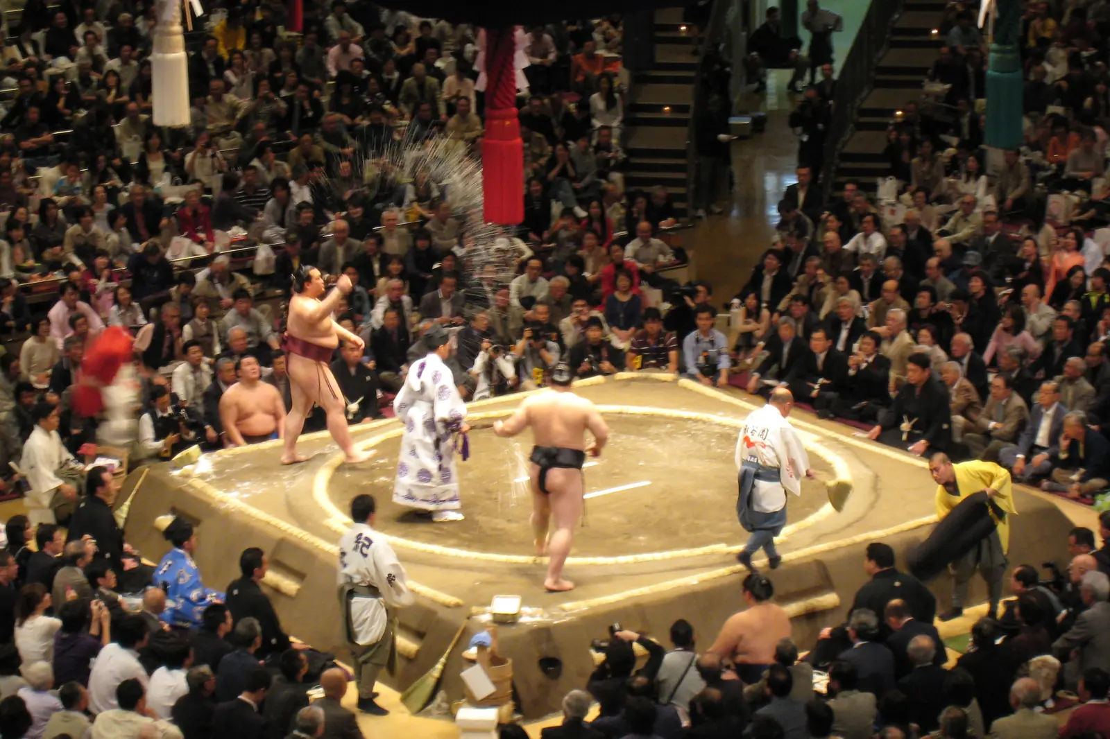 A wrestler throwing a handful of salt into a raised clay ring inside a packed arena, with a referee in white and other wrestlers waiting beside the ring